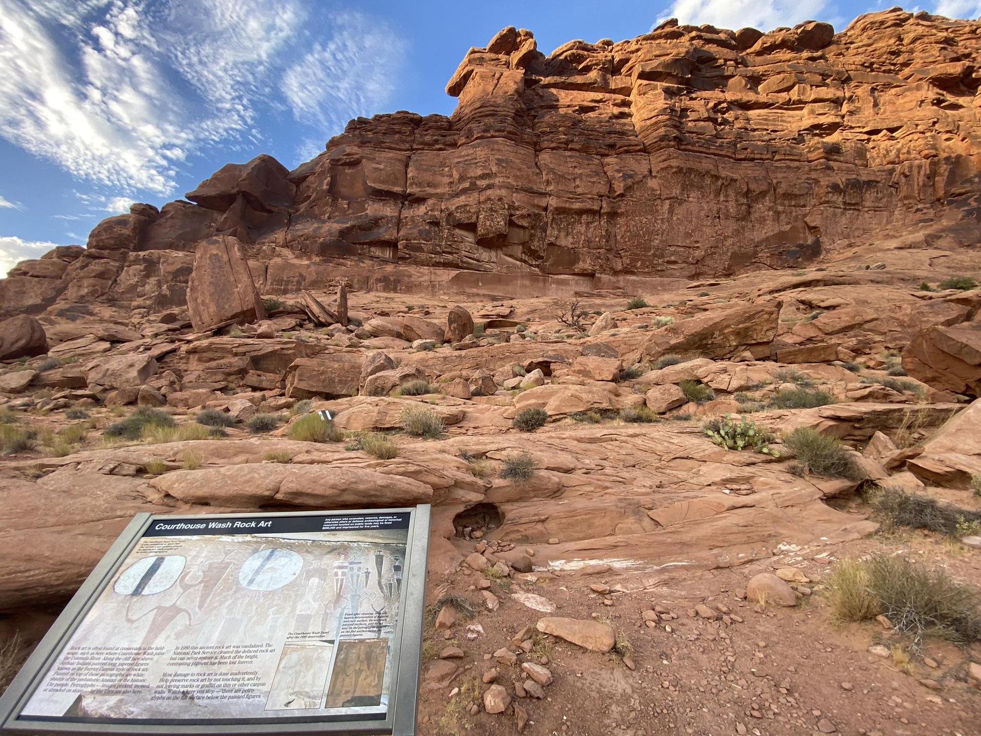 Information sign, large red rock cliff face, scattered rocks, sunny.