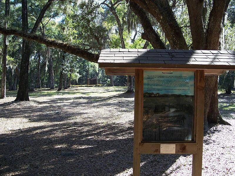 A wooden information sign in a sun-dappled forest with many trees.