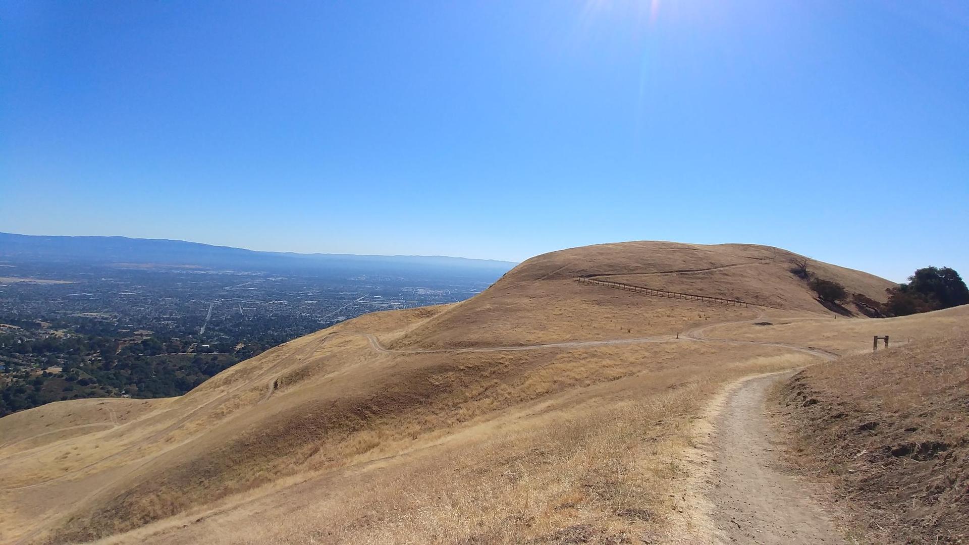 Dry, golden hills with a dirt path overlooking a distant city under bright sun.