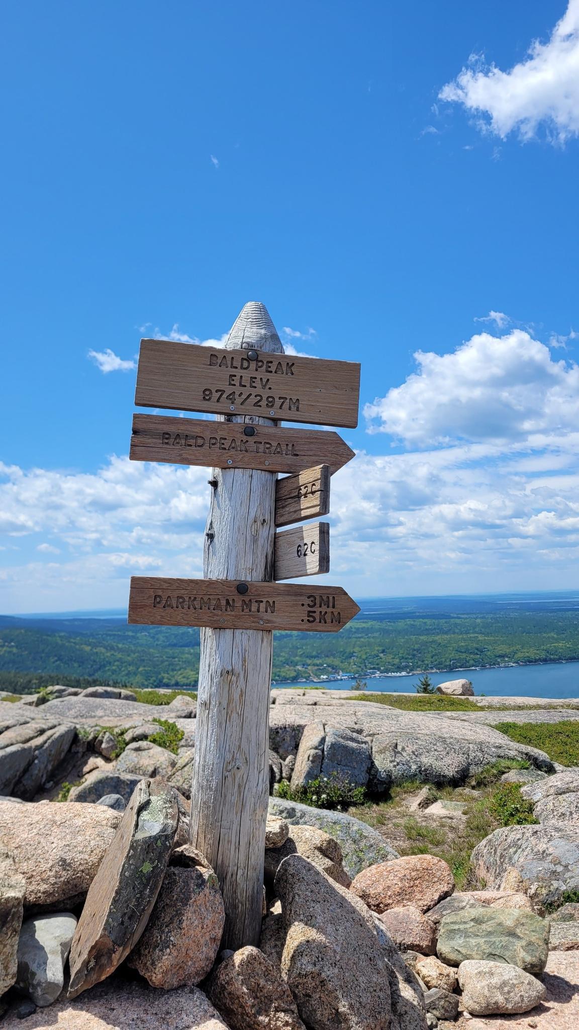 Wooden trail signpost on a rocky summit under a bright blue sky.