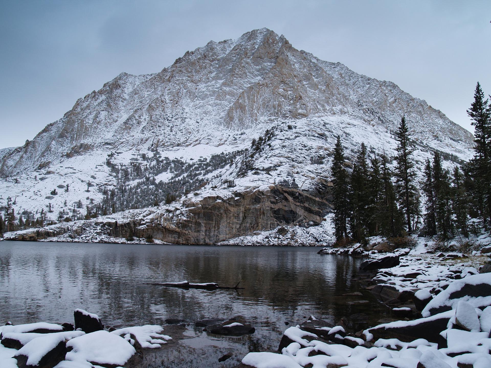 Snow-covered mountain overlooks a calm lake with pine trees on a cloudy day.