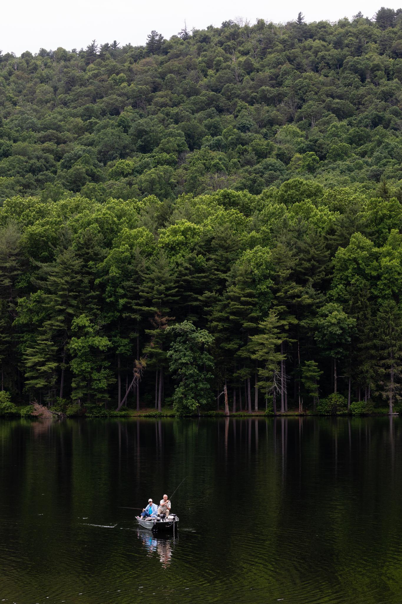 Two people fishing in a small boat on a calm lake, dense forest.