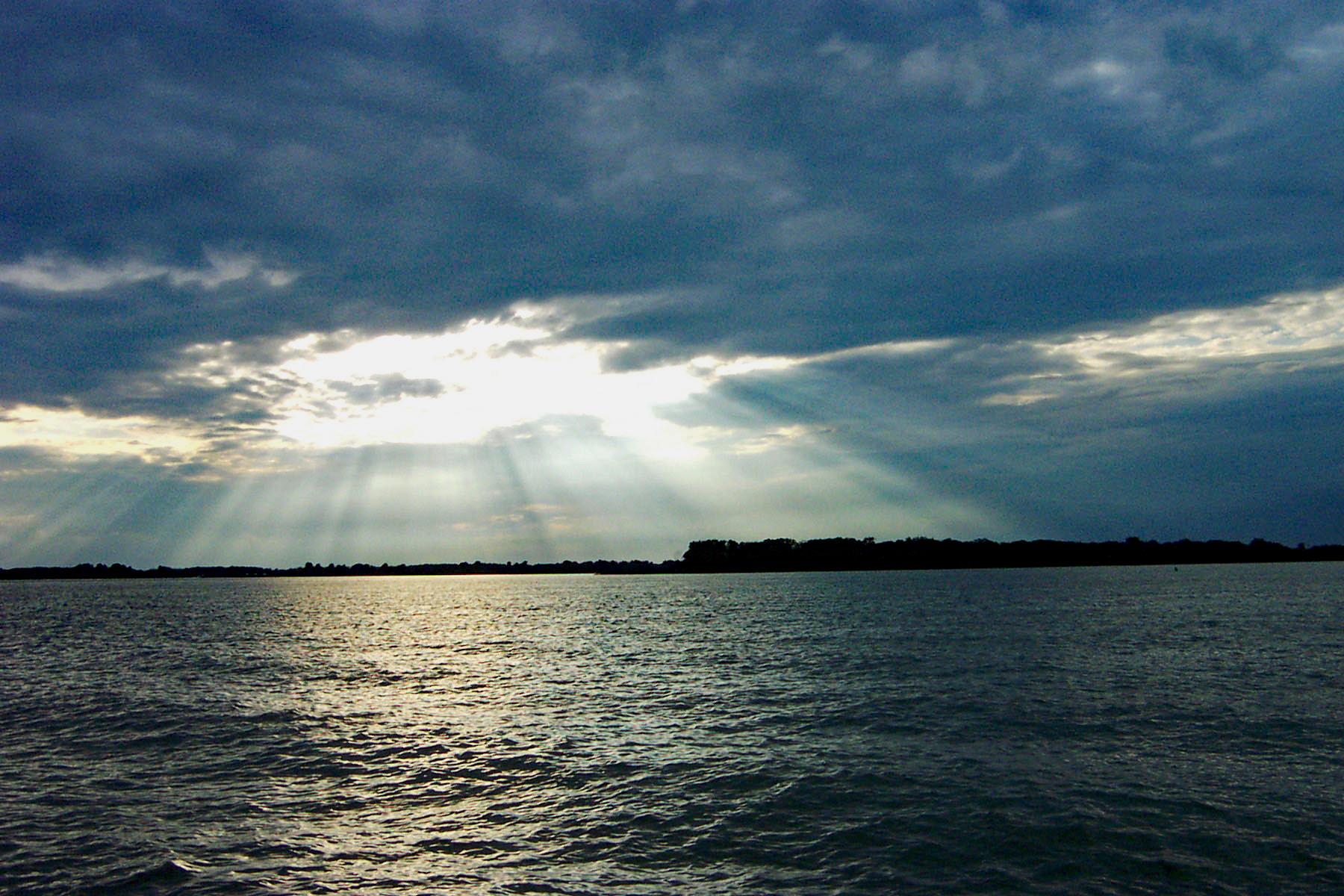 Sun rays pierce dark clouds over vast water, illuminating distant land.