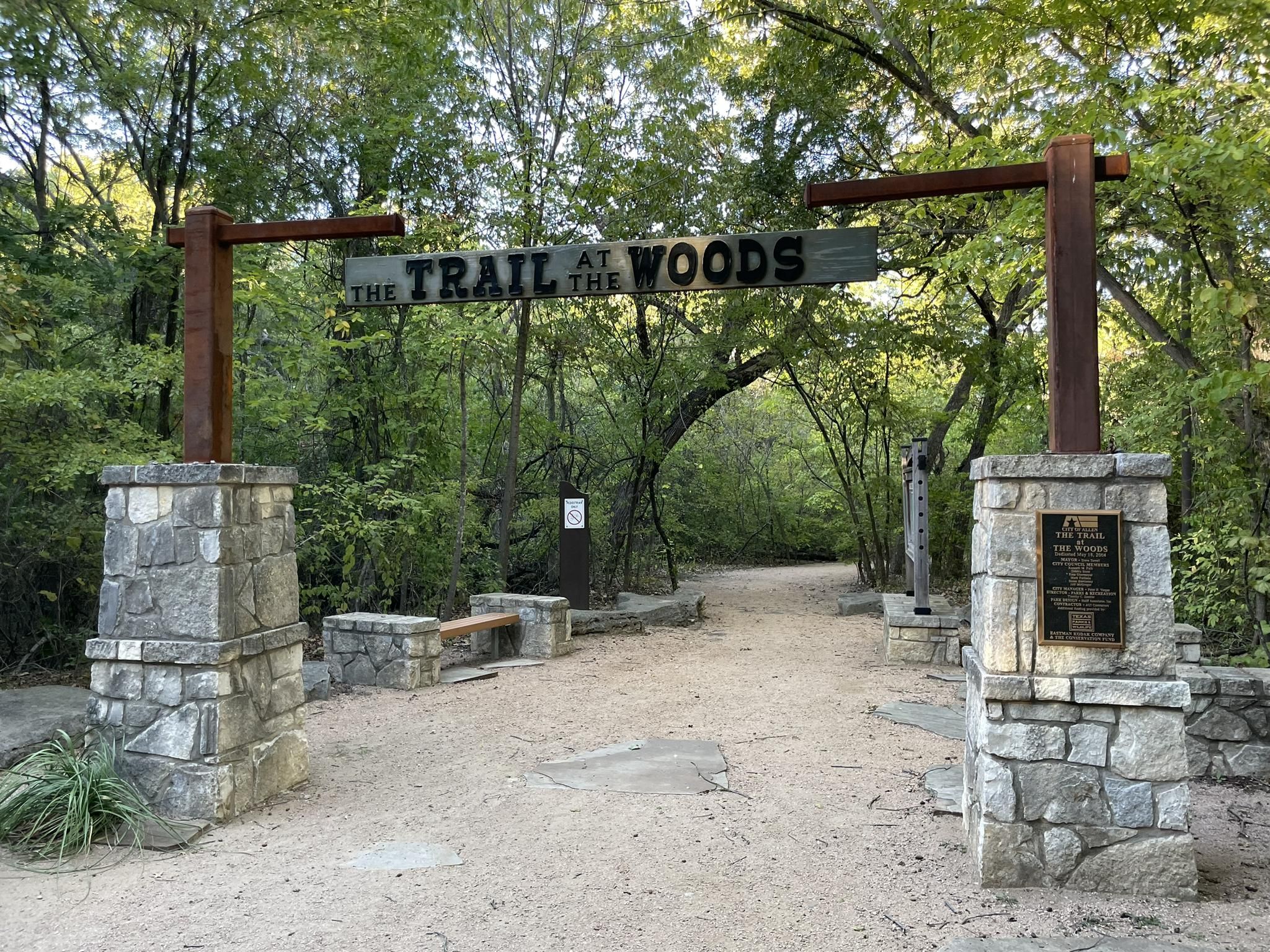 Wood and stone archway over a dirt path entering a green forest. - Pecan Grove Trail