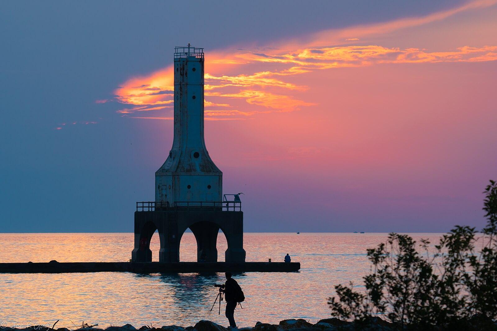A person photographs a lighthouse at sunset with a colorful sky over water.