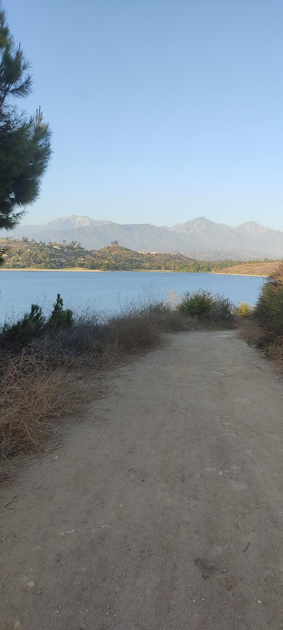 Dirt path overlooking a calm lake with distant mountains under a clear sky.