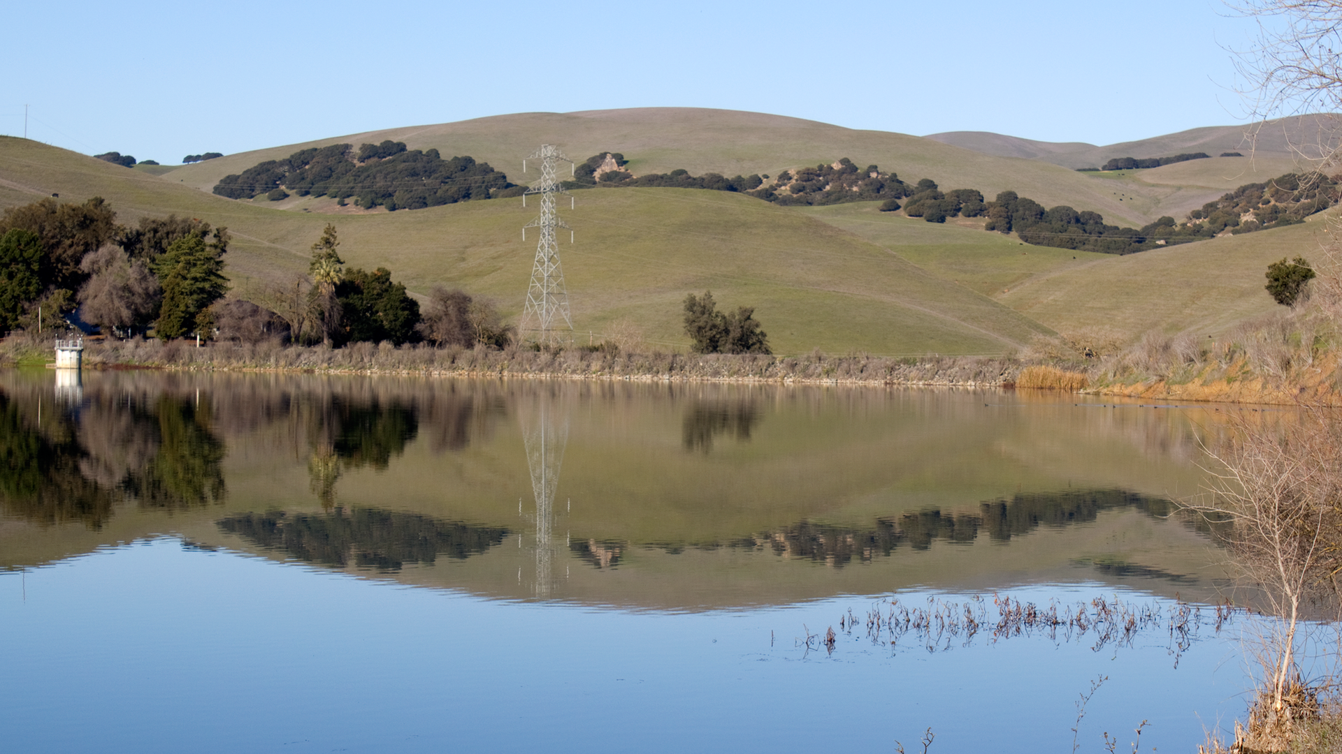 Calm lake reflecting green hills and a power line on a sunny day.