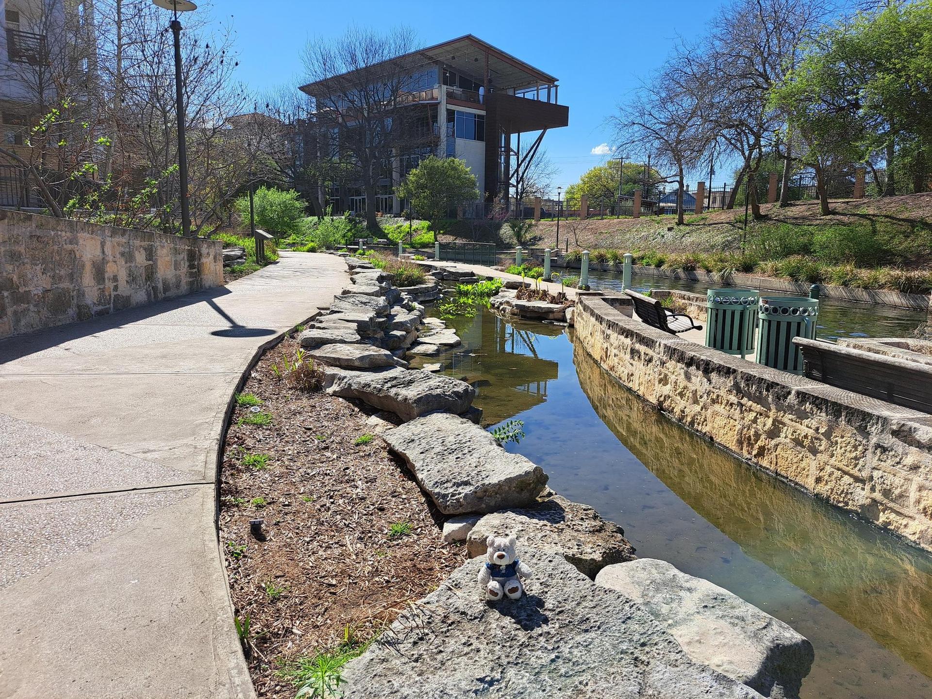 A small stuffed animal sits by a river with a modern building.