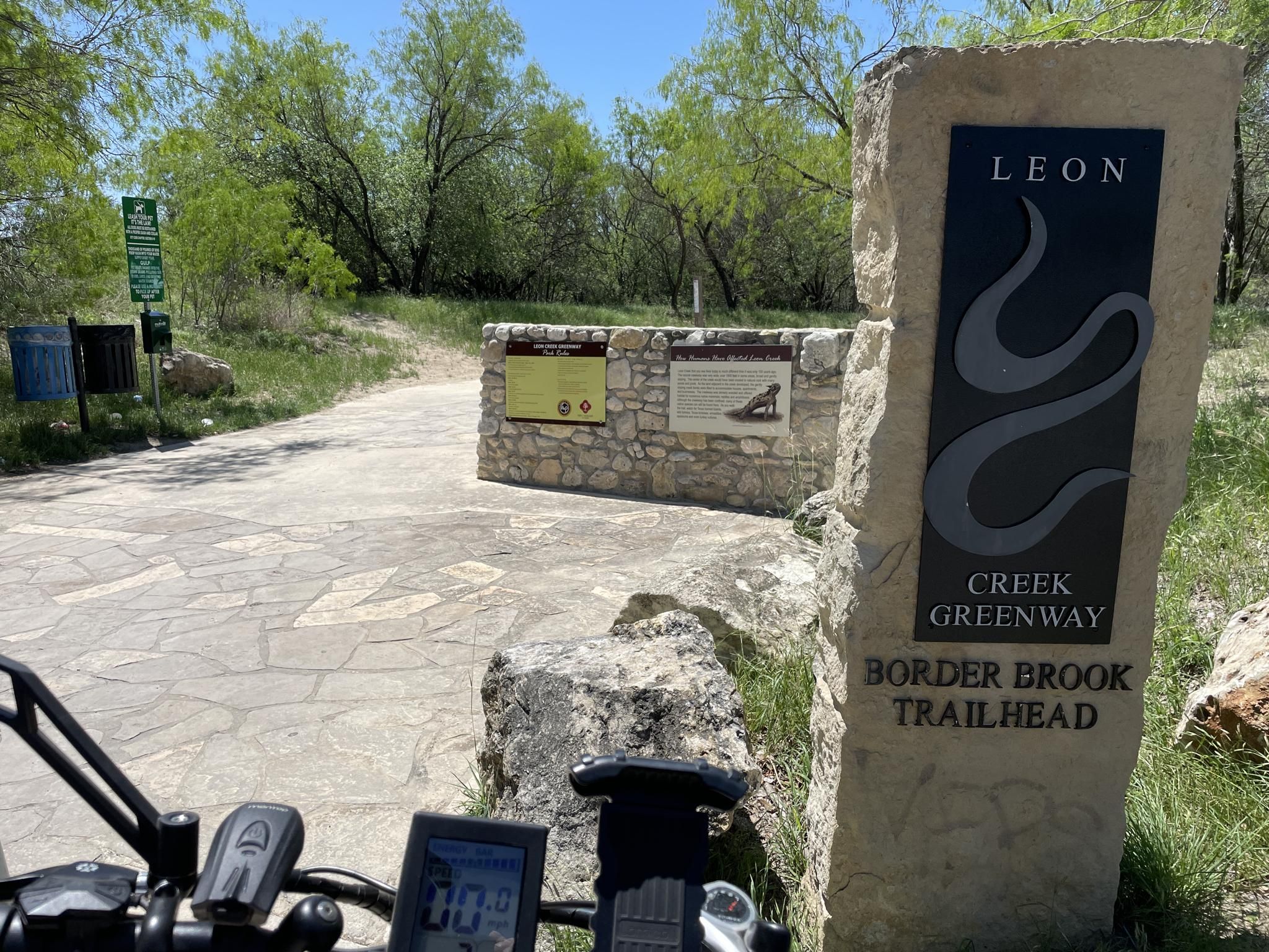 Stone trailhead sign and information boards on a sunny day, with a bike. - Leon Creek Greenway Trail