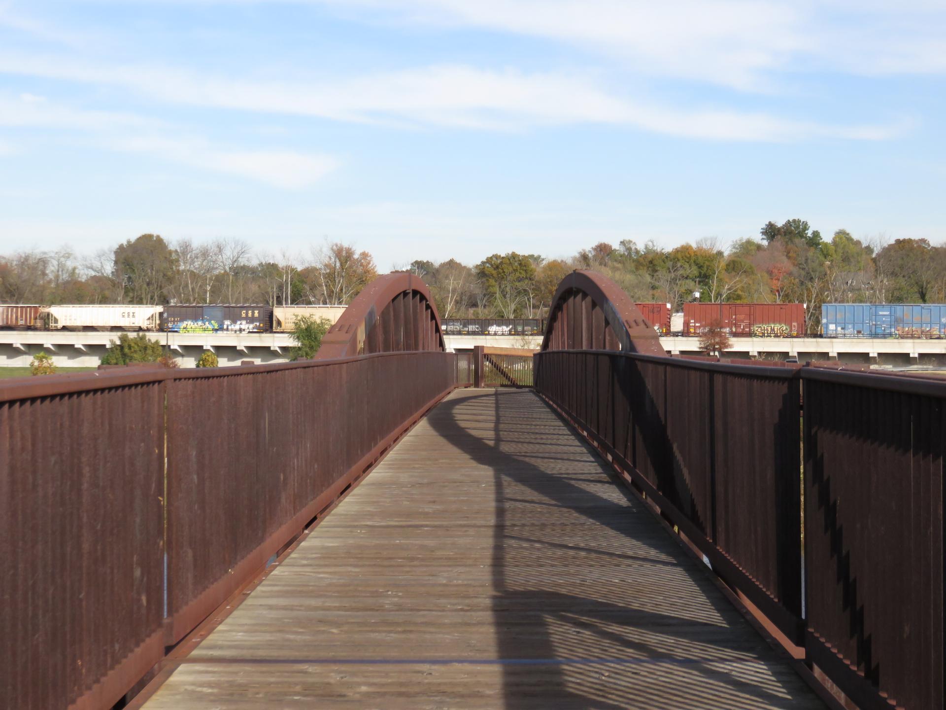 A wooden bridge with arched metal railings over a train track under a blue sky.