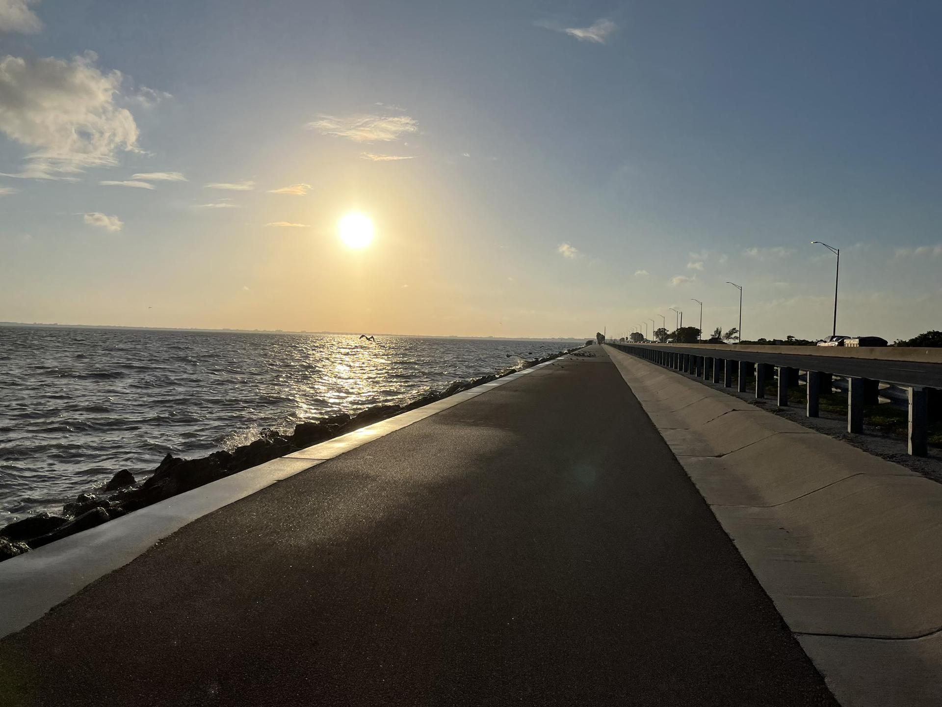 Paved path alongside ocean at sunset, with a bridge in the distance.