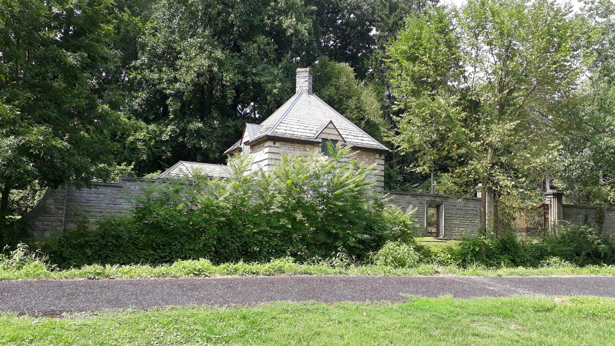 Stone house with a tall wooden fence, surrounded by green trees.