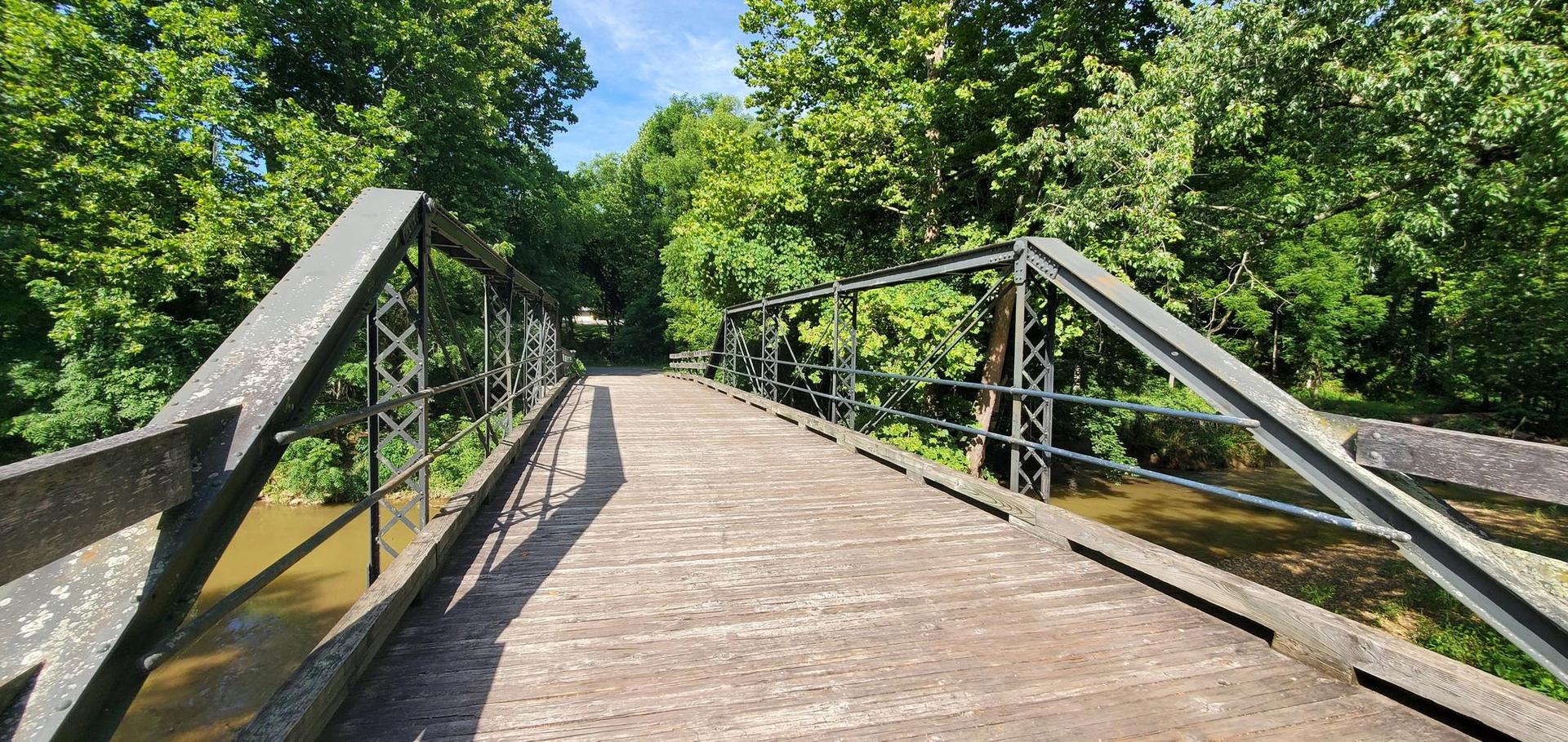Wooden and metal bridge over a river, surrounded by lush green trees on a sunny day.