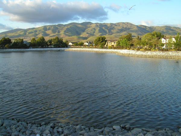 Calm lake with rocky shores, houses, and mountains under a cloudy sky.