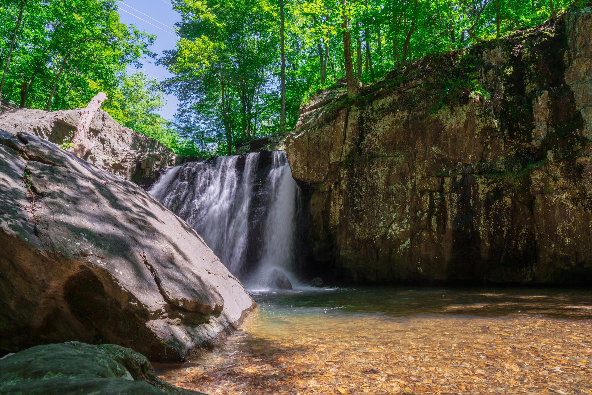 A waterfall cascades into a clear pool surrounded by lush green forest and large rocks.