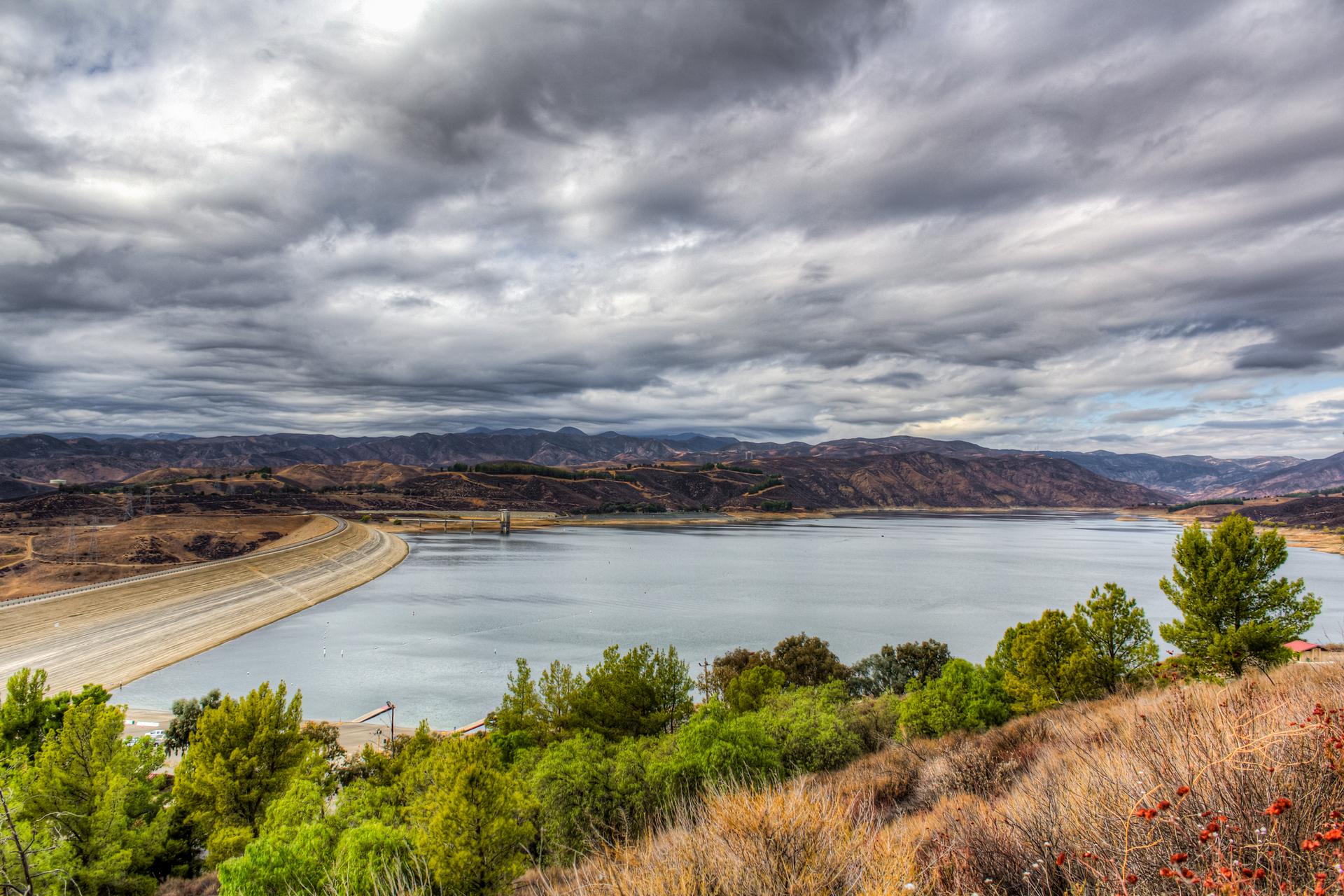 Expansive lake and dam under a dramatic, cloudy sky with green trees.