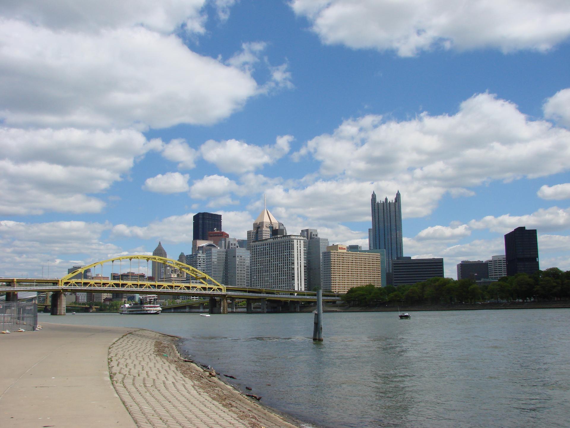 A city skyline with a yellow bridge over a river on a bright, cloudy day.
