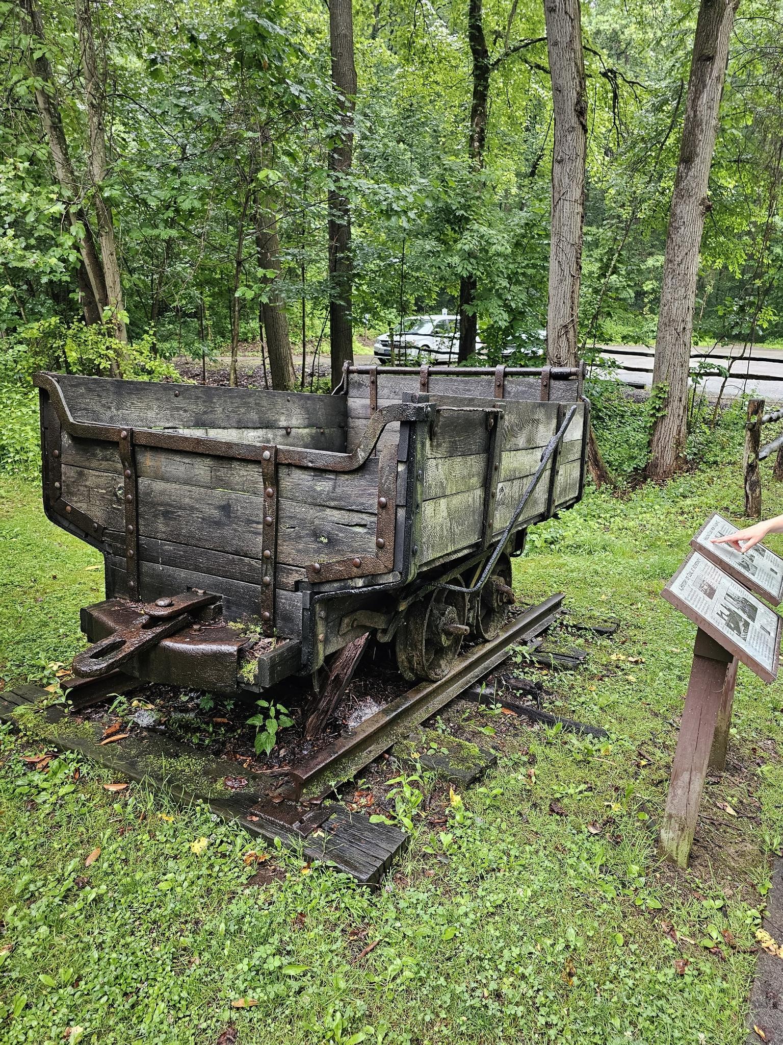 An old wooden mining cart on tracks in a forest, with a hand pointing at a sign.