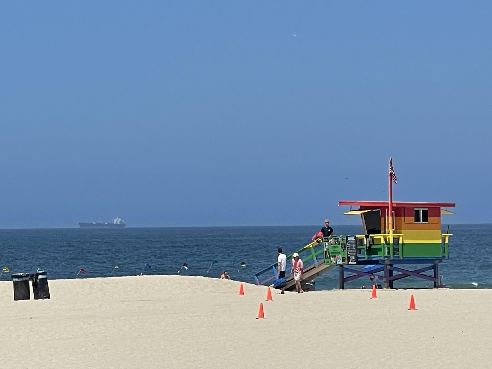 Rainbow lifeguard tower on a sunny beach with people and a distant ship.