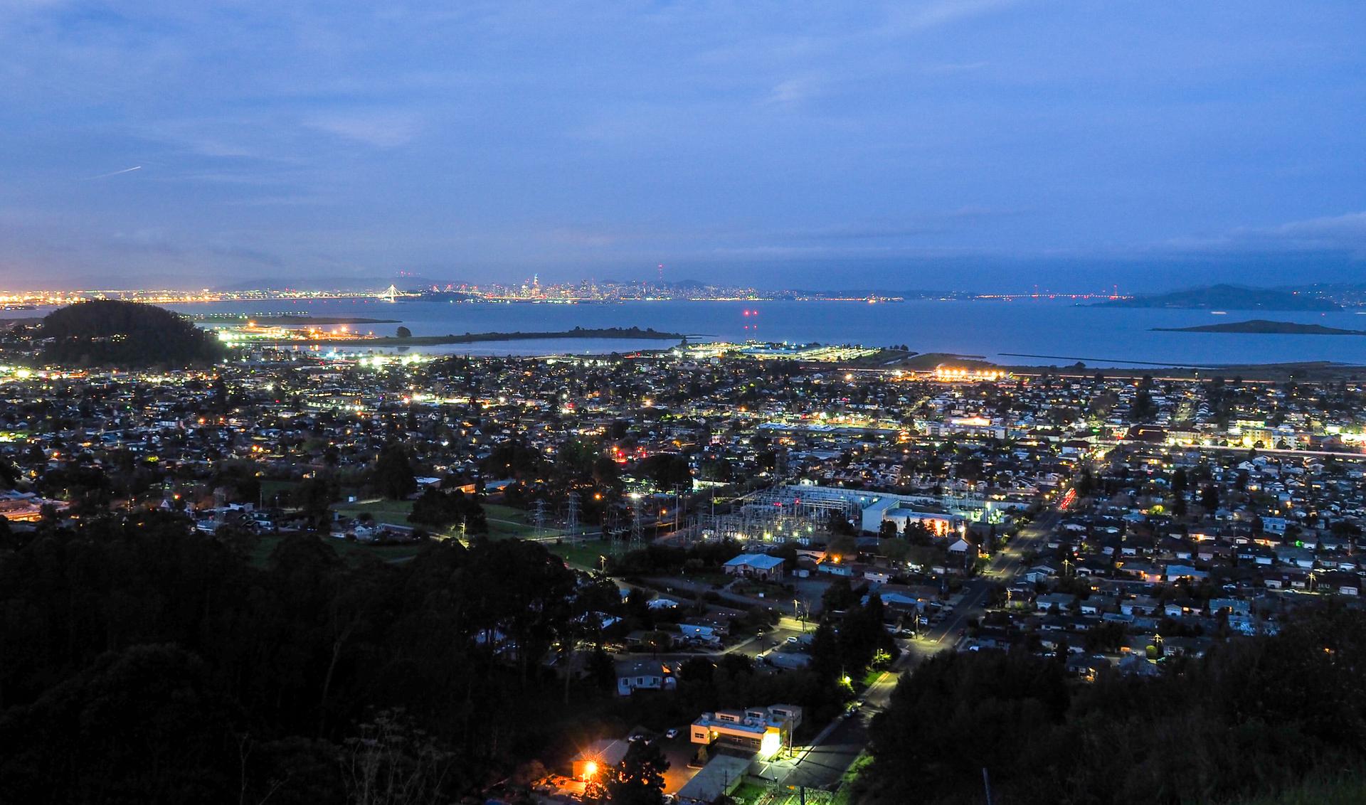 An illuminated city at dusk, with a bay and distant urban lights under a dark sky.
