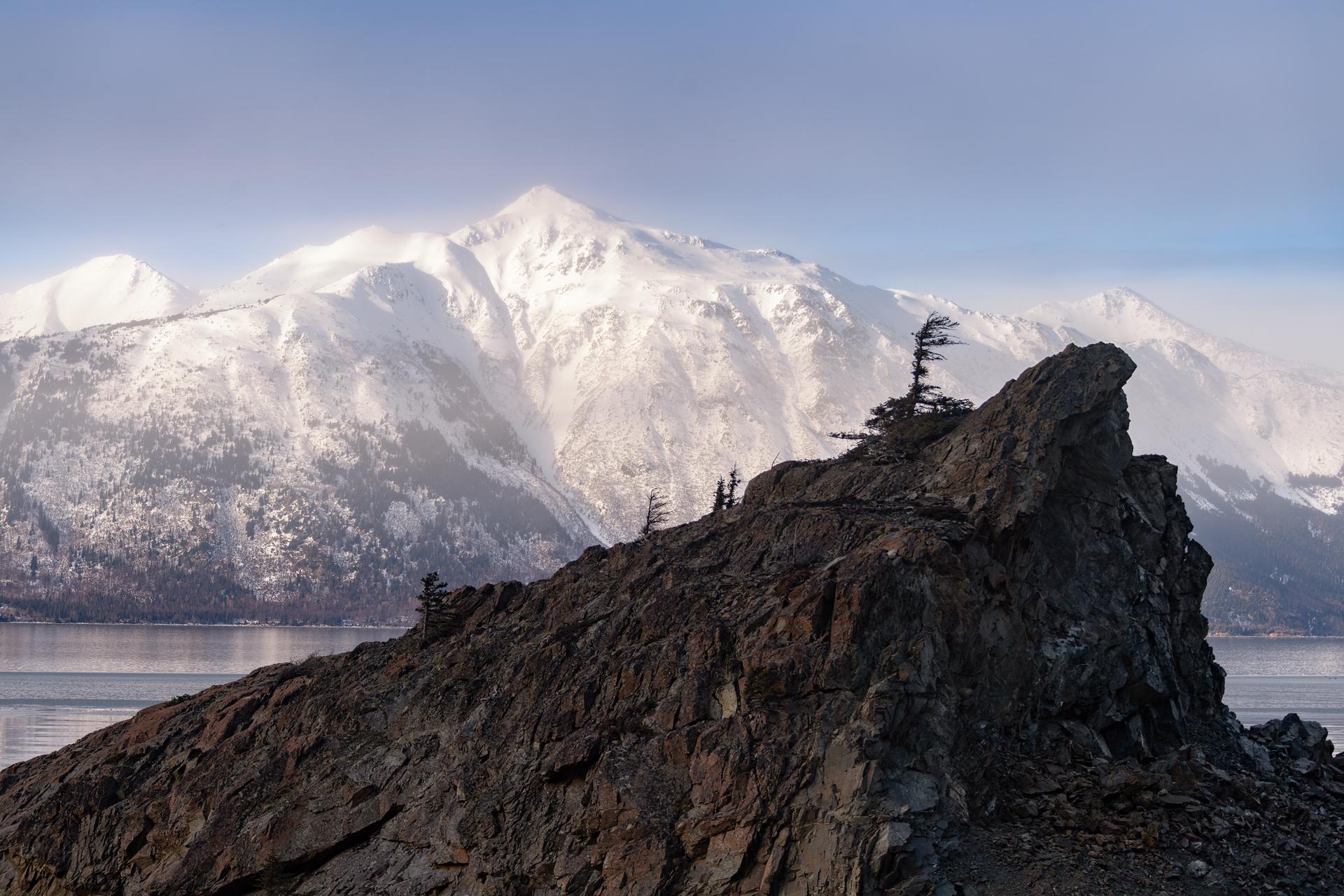 Hikers on a dark rock formation with a vast snowy mountain range and water.