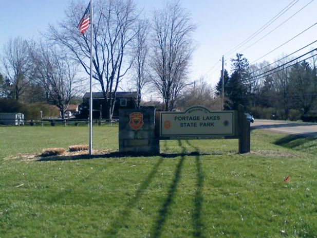 A park sign and flagpole stand on a sunny, grassy field with bare trees. - Shoreline Trail