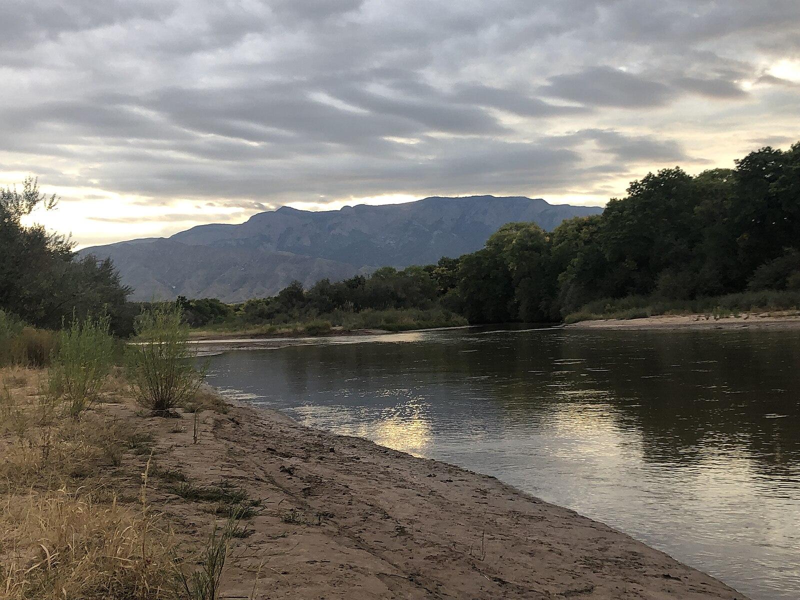River flows past a sandy bank with mountains under a cloudy sky.