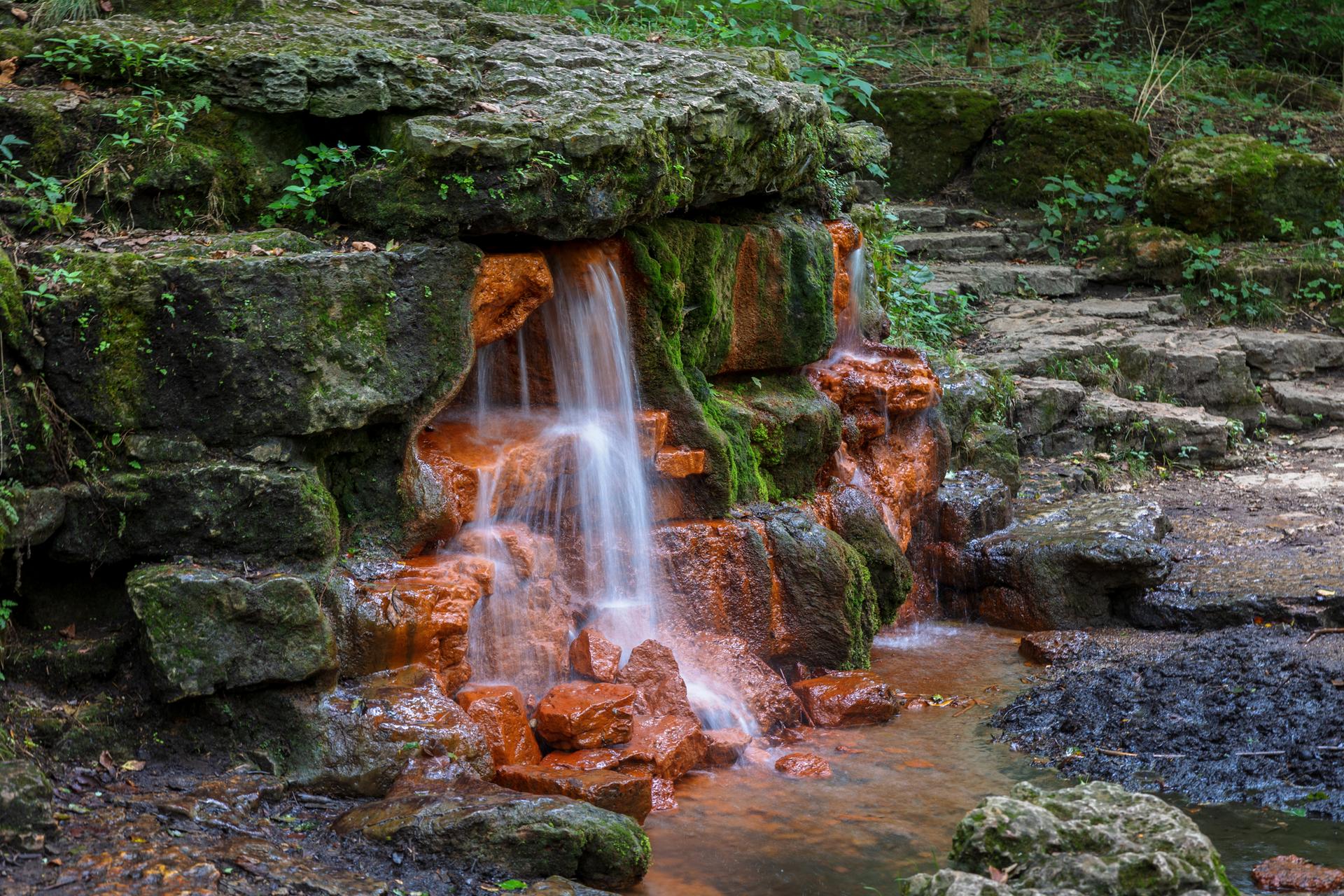 A small waterfall cascades over orange and mossy rocks into a stream.