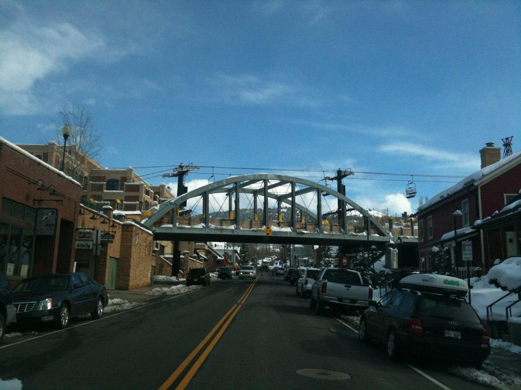 Sunny street with cars leading to an arched bridge, snow on buildings.