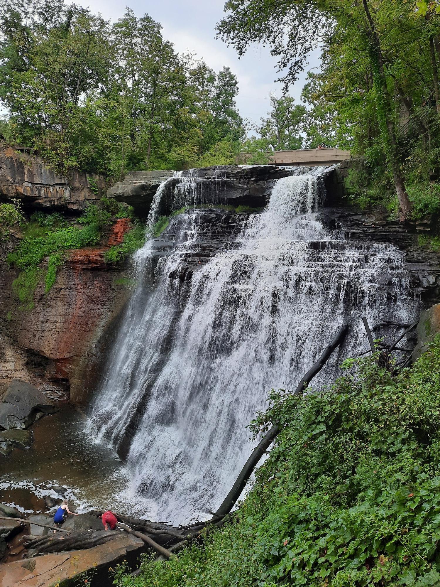 Large waterfall cascades over rocks into a pool, surrounded by green trees.