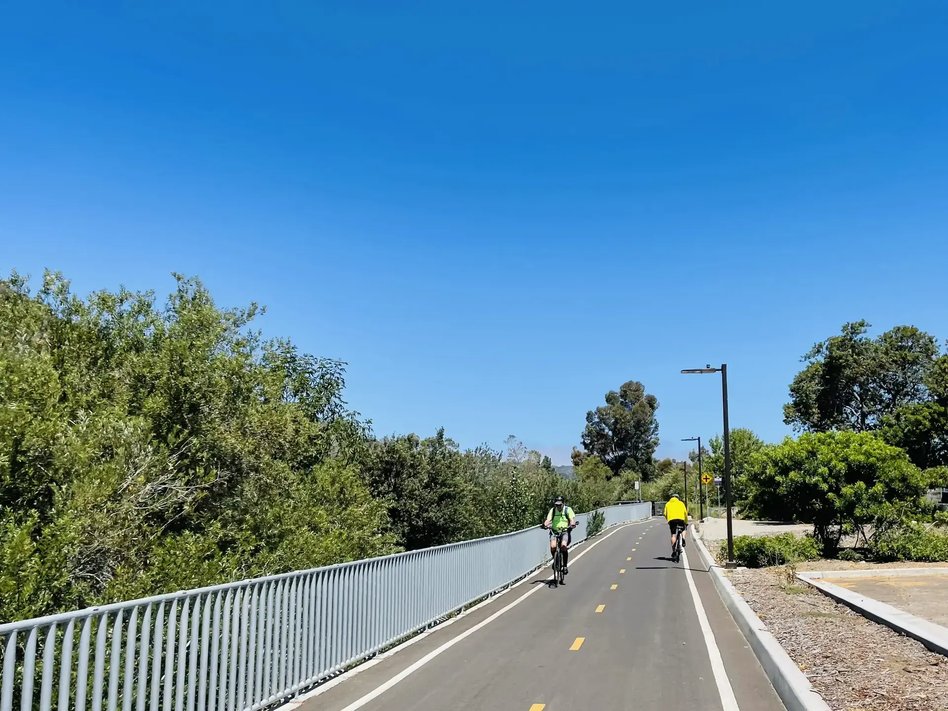 Two cyclists ride a paved path bordered by green trees under clear sky.