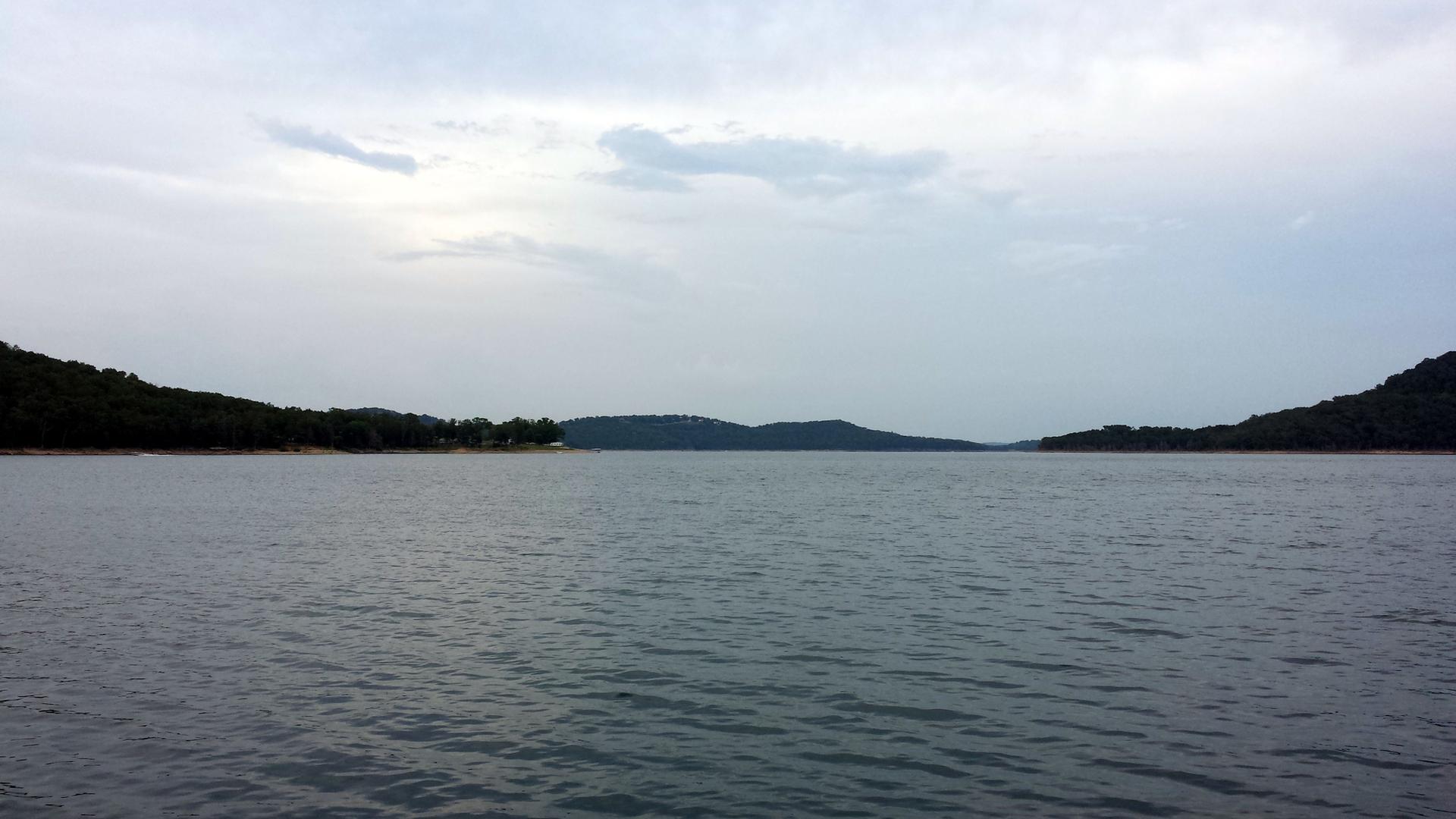 Wide view of a calm lake surrounded by forested hills under a cloudy sky.