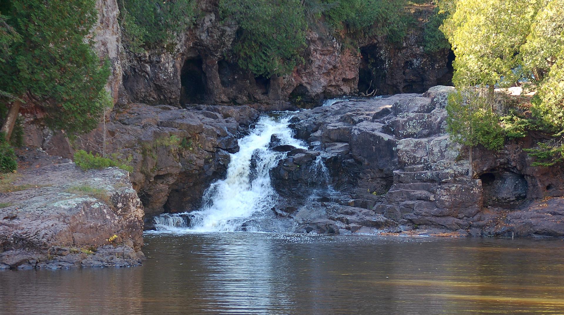 Waterfall cascades over dark rocks into a river, surrounded by cliffs and trees.