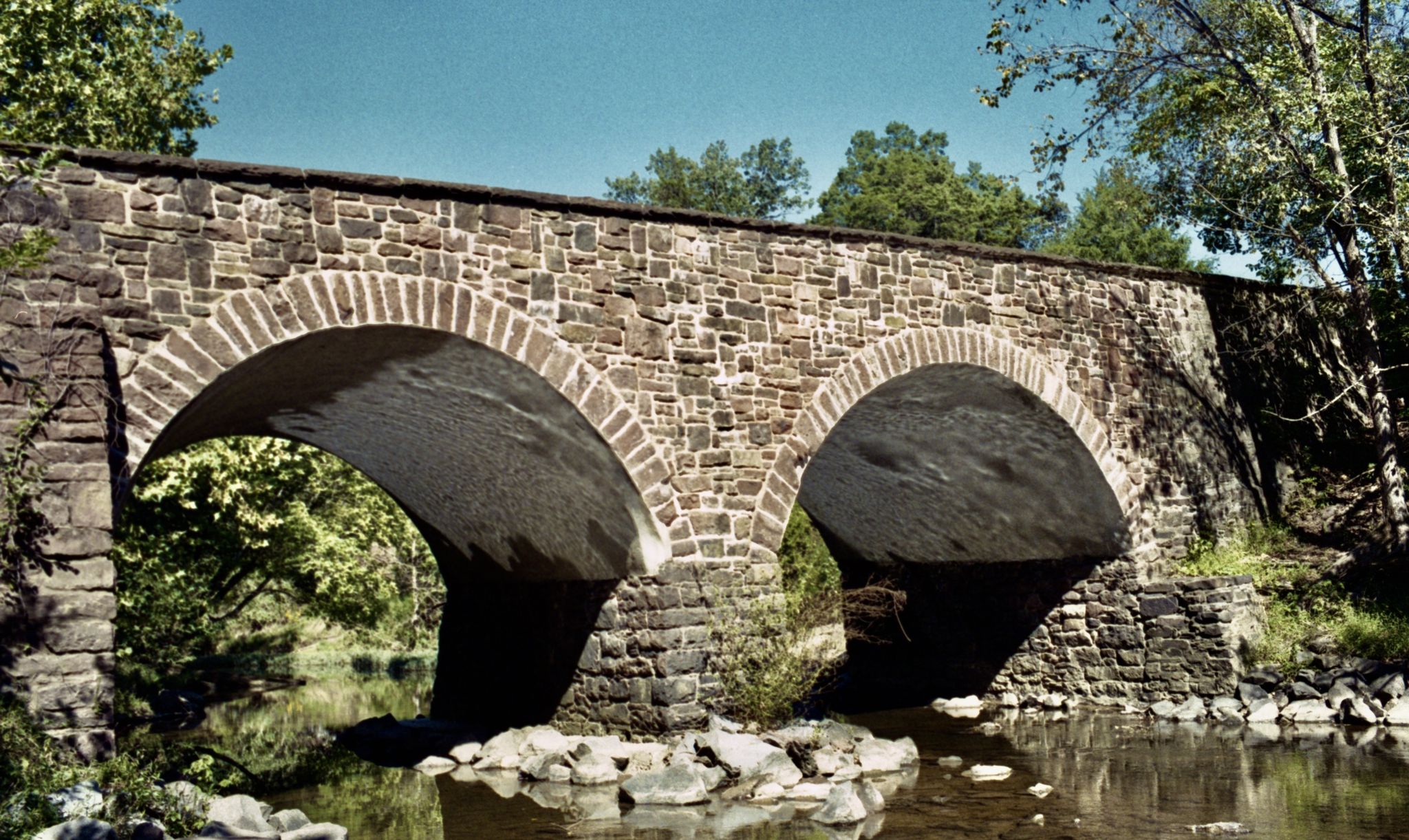 A stone arch bridge spans a calm stream under a clear sky. - Bull Run & Manassas National Battlefield Park loop