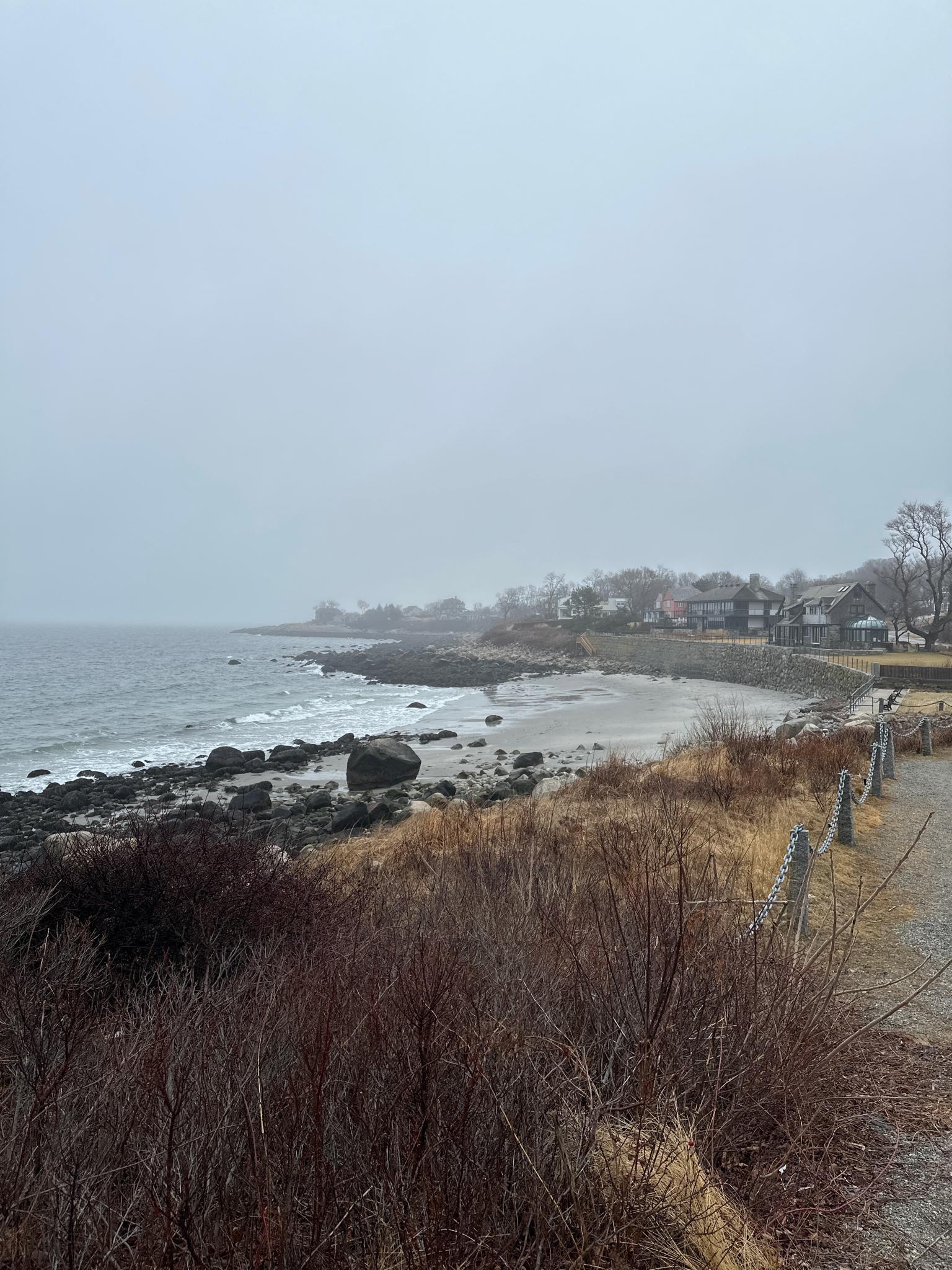 Overcast day on a rocky coastline with a sandy beach and distant houses.