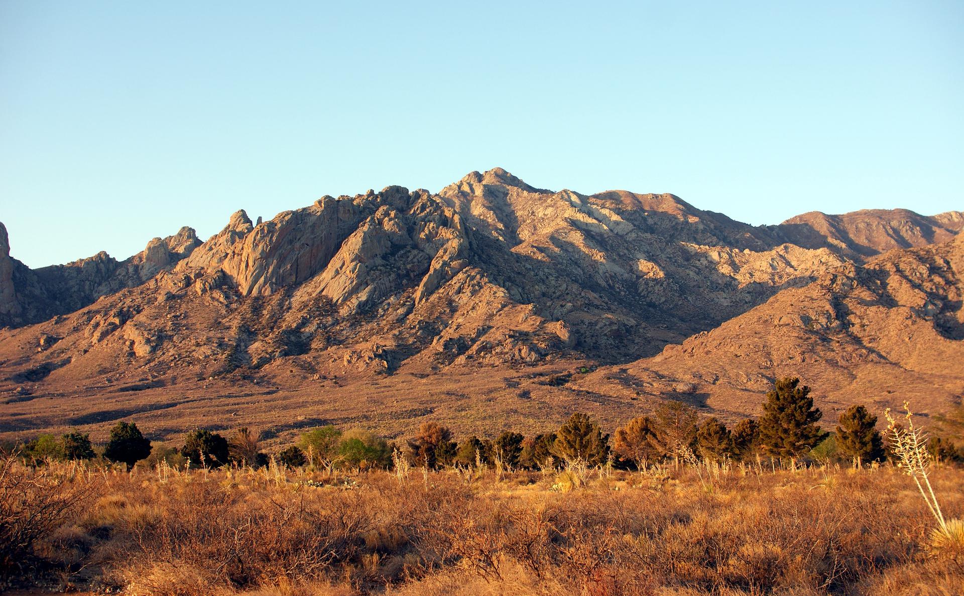 Rugged mountains glow in golden light over a dry, shrub-filled landscape.