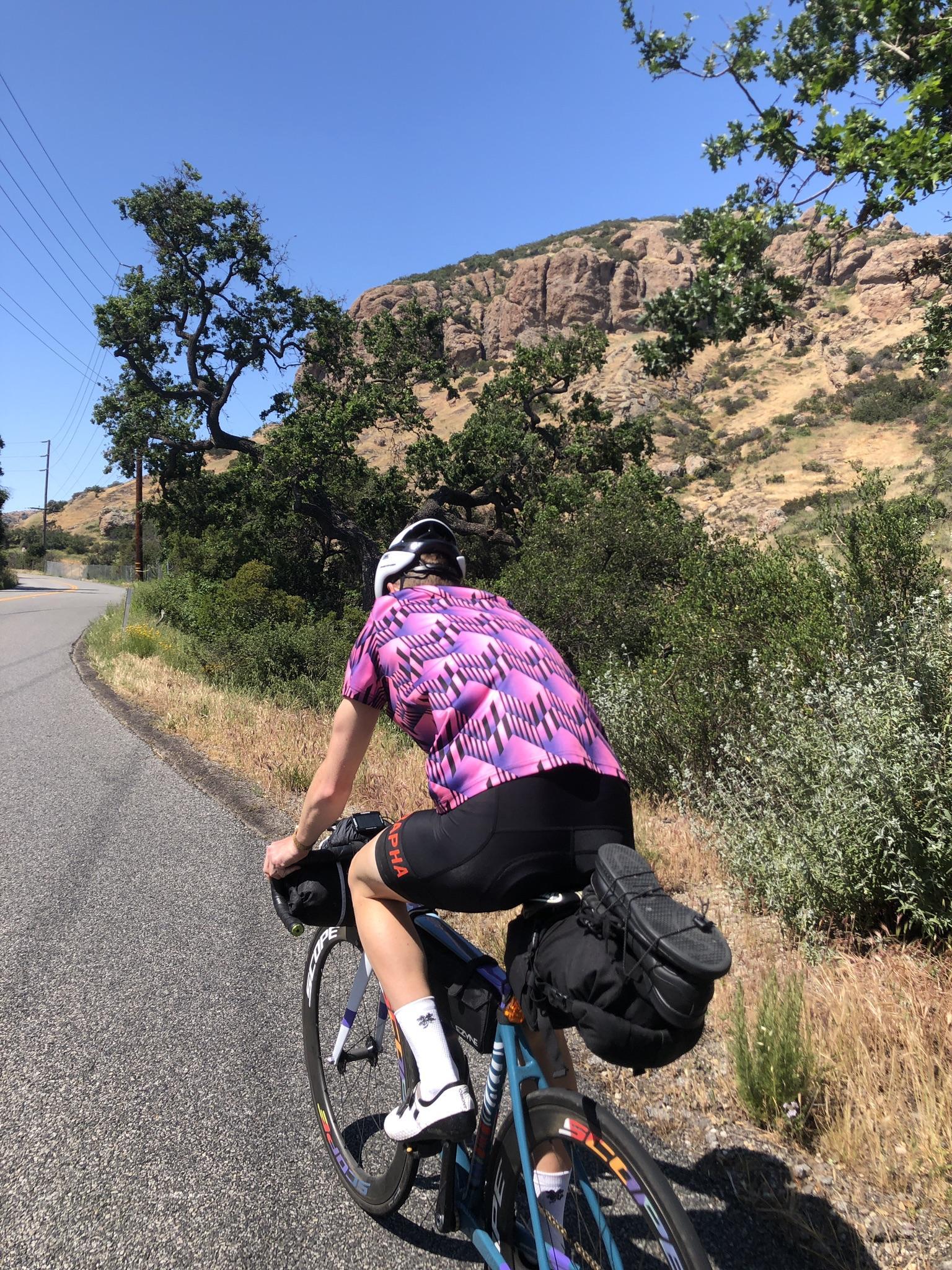 Cyclist on a paved road, with rocky hills and green trees under bright sun.