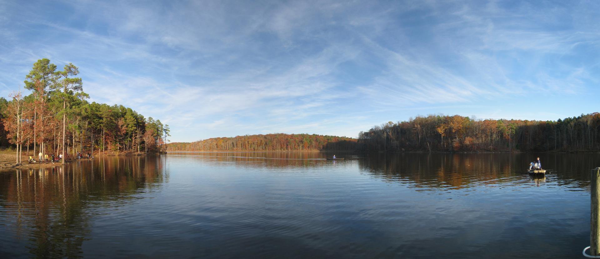 Panoramic lake view with autumn trees, people on shore, and a boat.