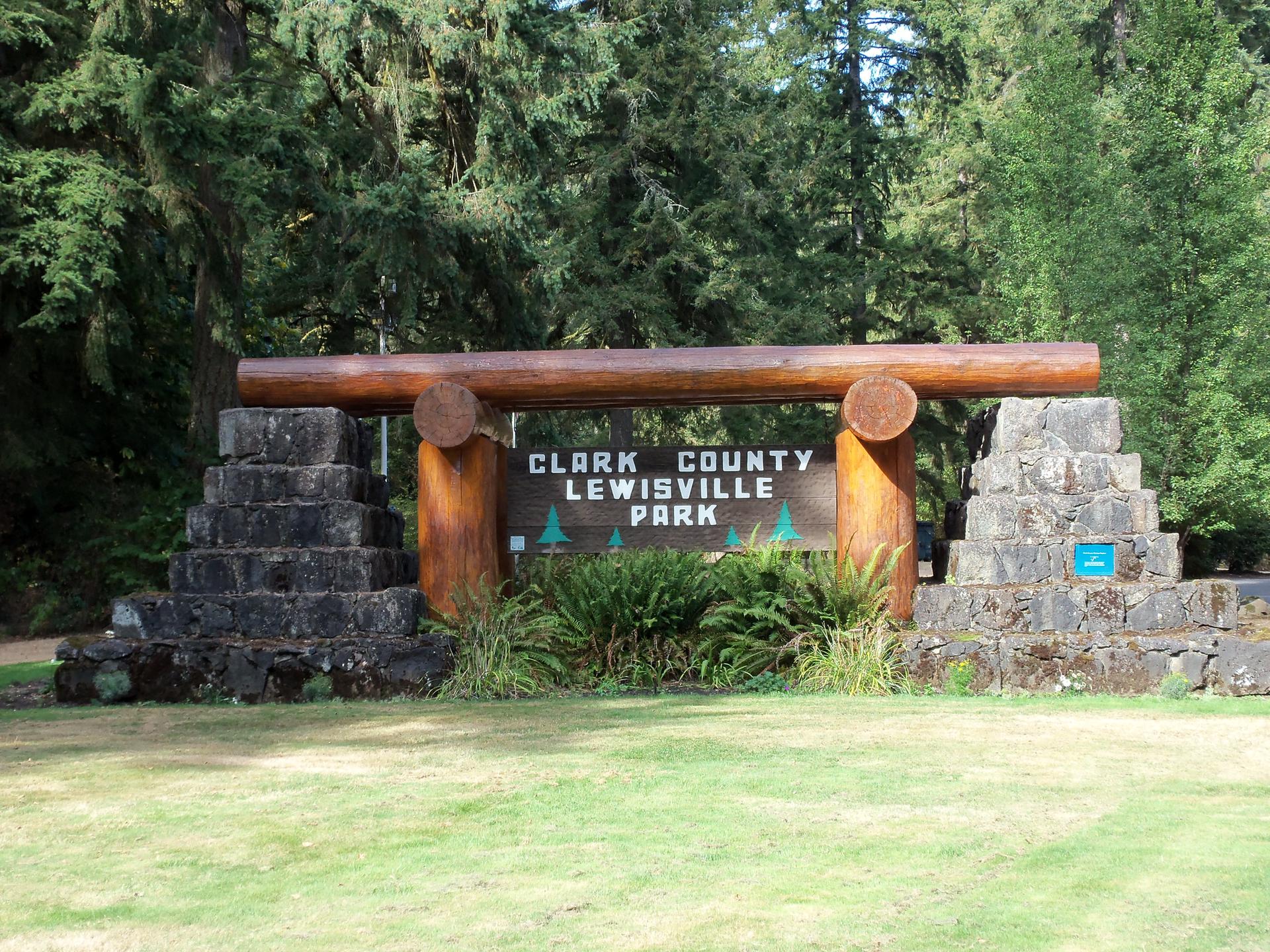 Sunny view of a large wooden park sign surrounded by grass and tall trees.