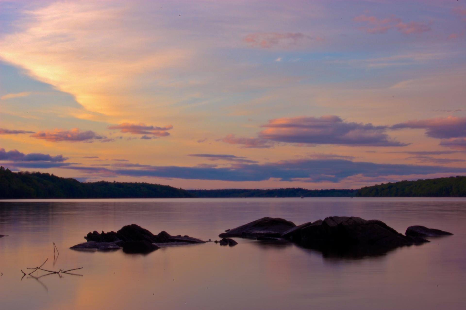 Serene lake at sunset with colorful sky, dark rocks, and forested shoreline.