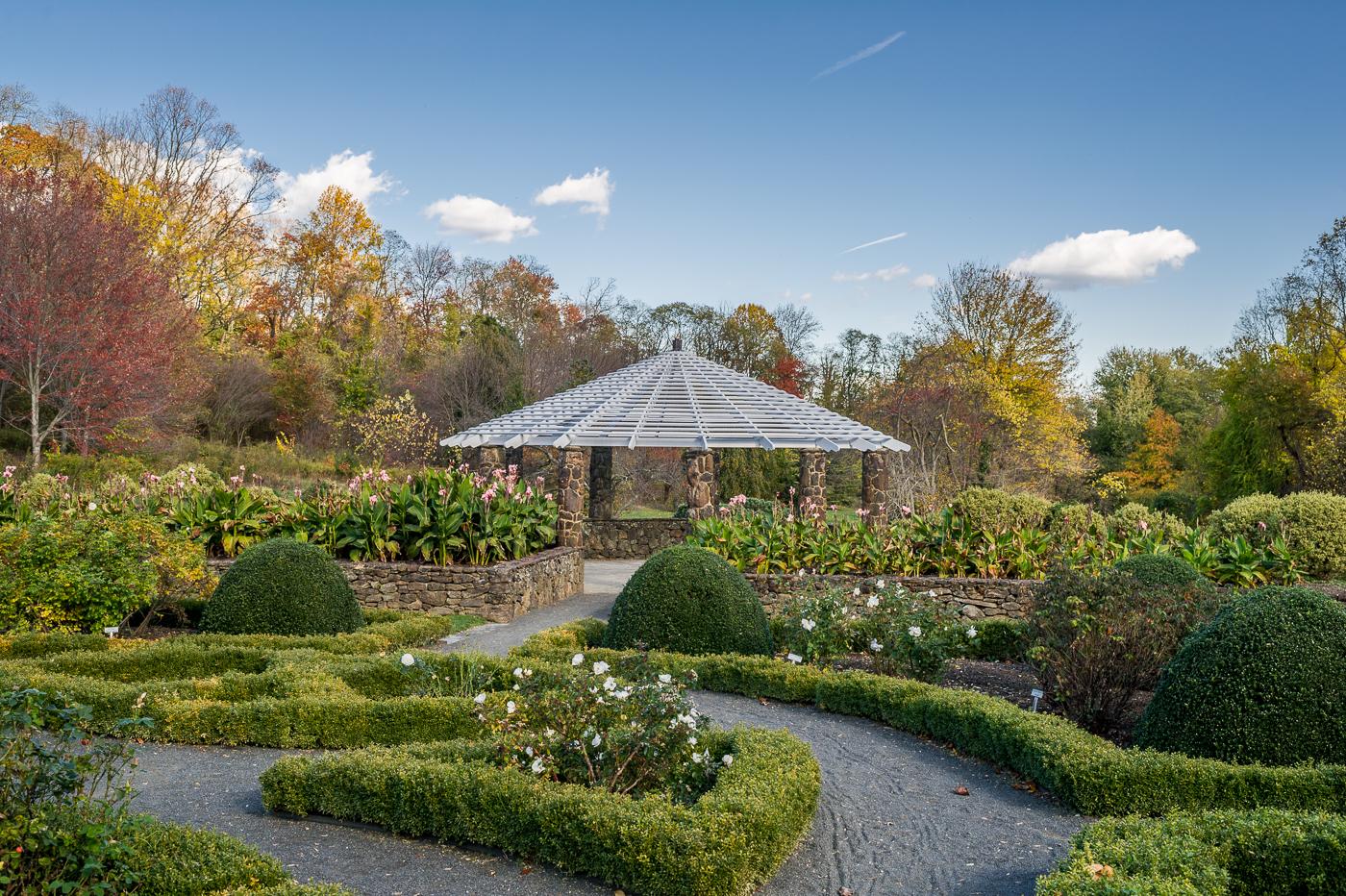 A stone gazebo surrounded by manicured hedges and autumn trees under a blue sky.