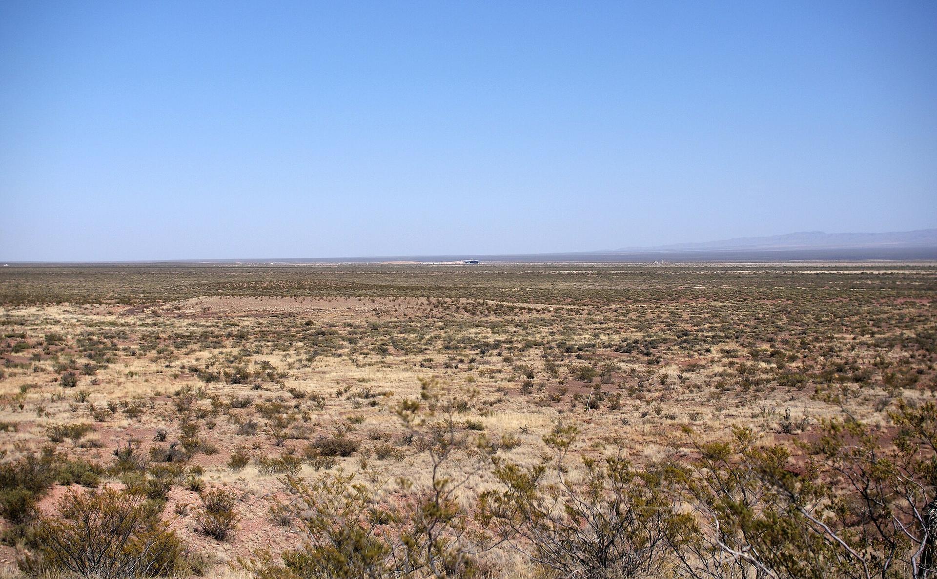 Vast, sunlit desert landscape with sparse scrub under a clear blue sky.