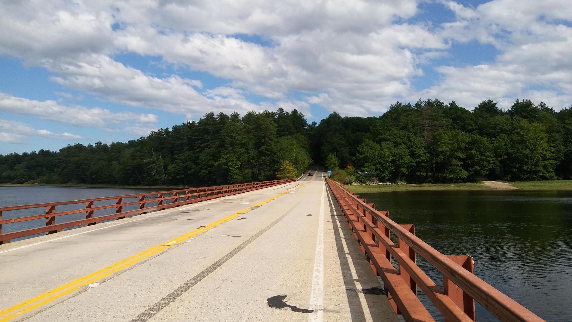 A long bridge crosses water towards a forest under a bright, cloudy sky.