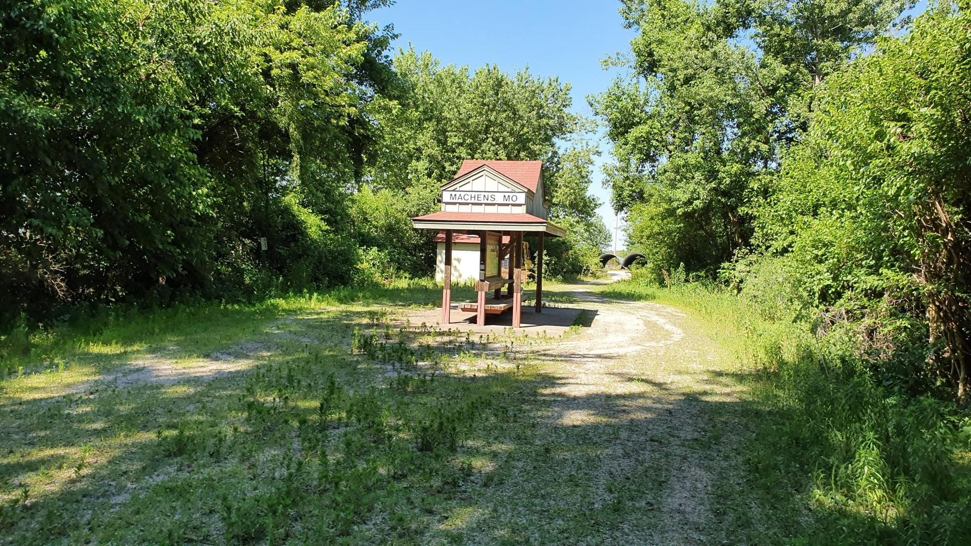 Sunny, grassy path leading to a small, red-roofed building amidst green foliage.