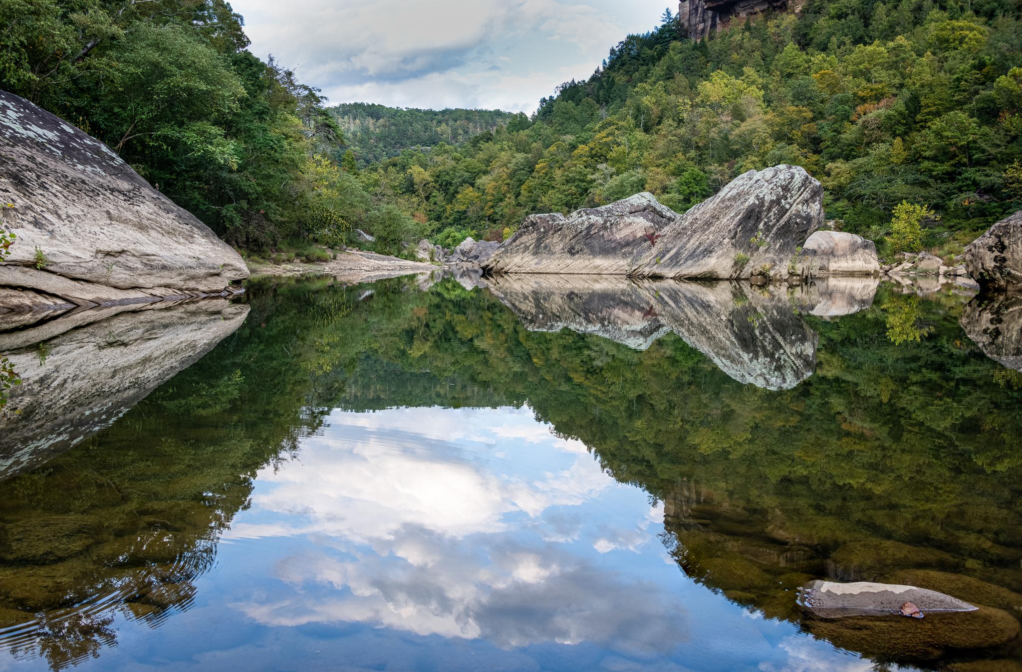 Calm river reflecting sky and green forest, flanked by large rock formations.