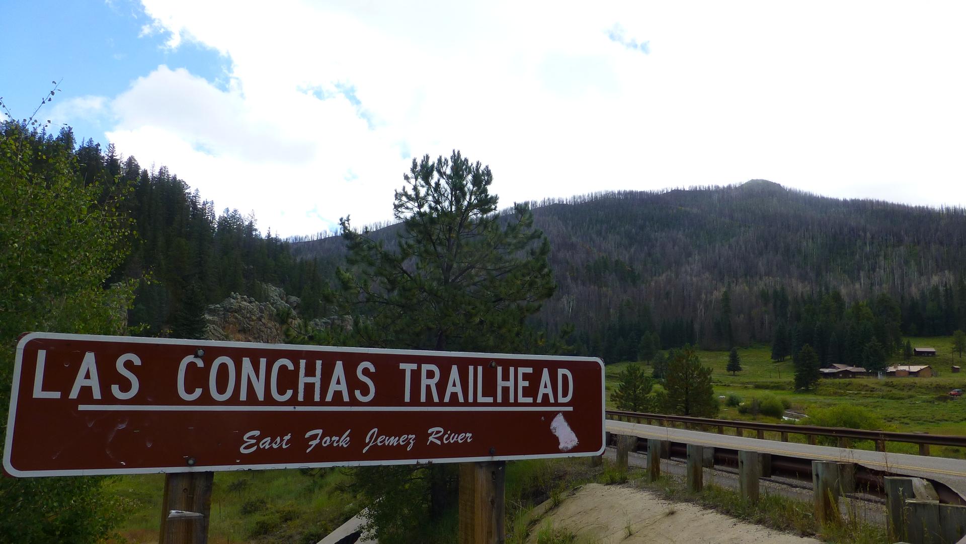 A brown sign marks a trailhead by a bridge, with forested hills beyond.