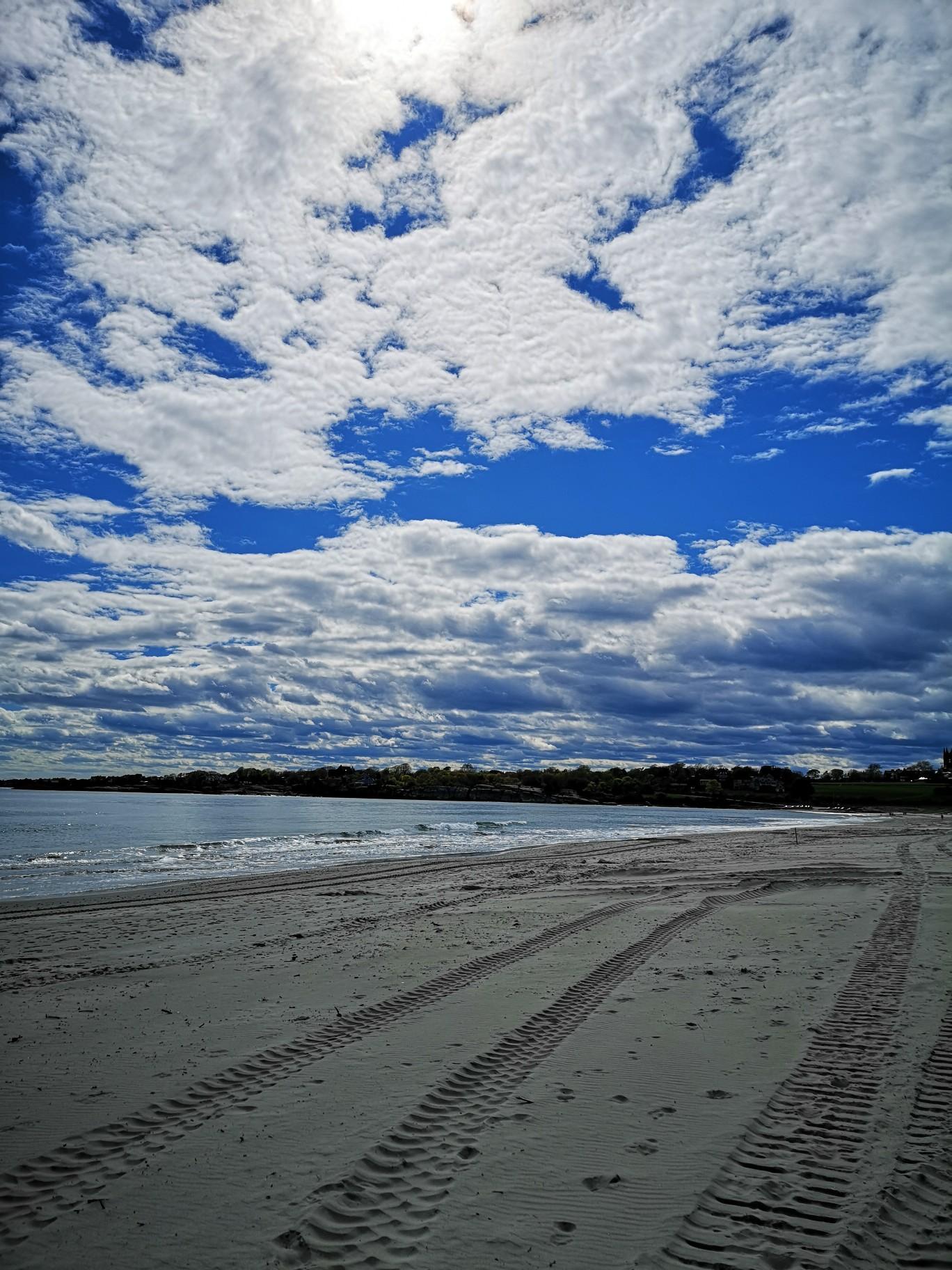Sandy beach with tire tracks under a bright blue sky with fluffy clouds.