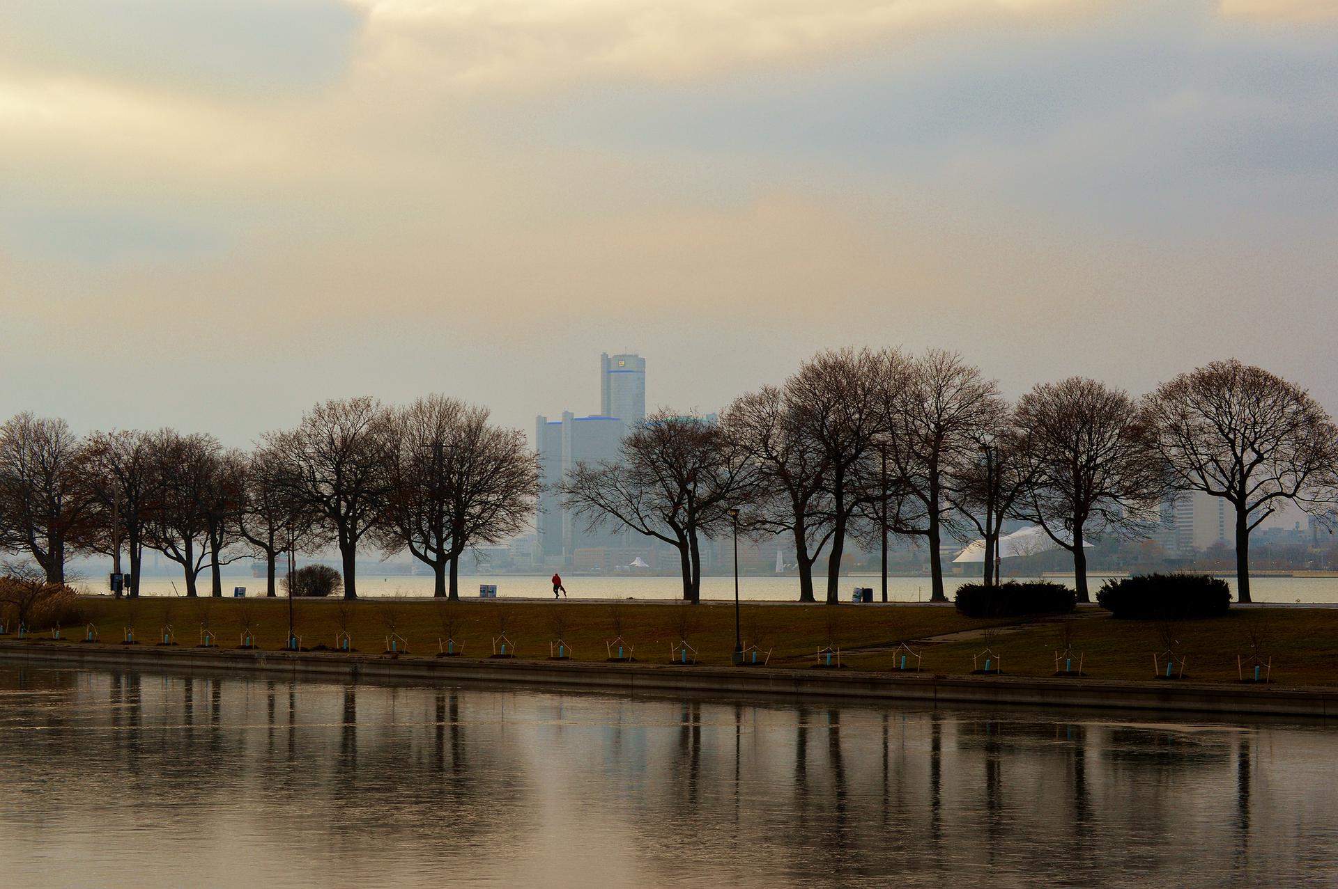 Bare trees line a waterfront path with a distant city skyline.