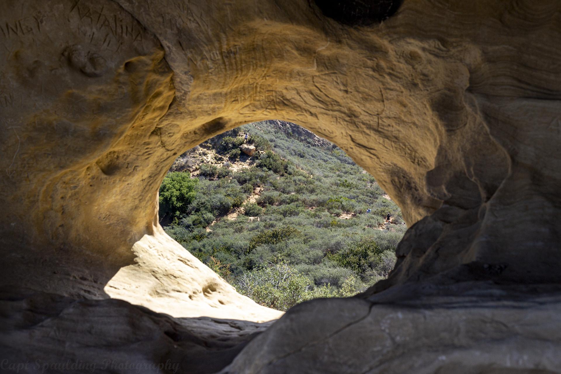 View through a natural rock arch to green, sunlit hills.