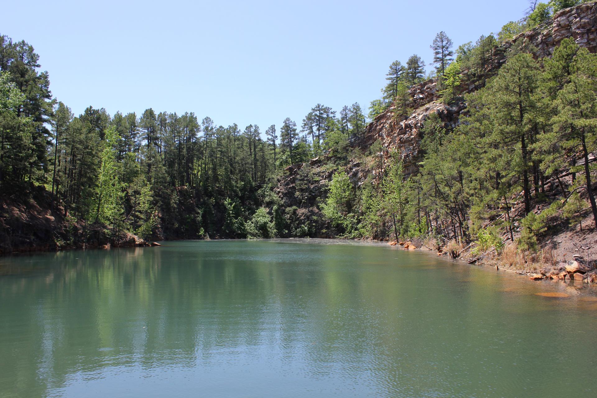 Calm green lake surrounded by steep, rocky, pine-covered hills on a sunny day.